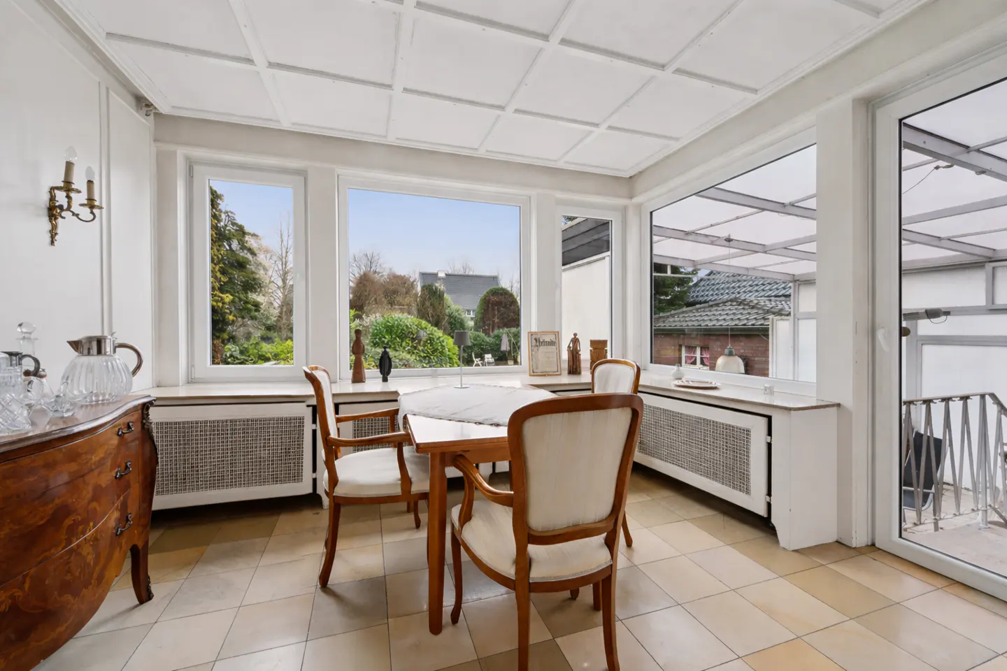 Bright sunroom with a wooden table and chairs, large windows overlooking a garden, and a tiled floor.
