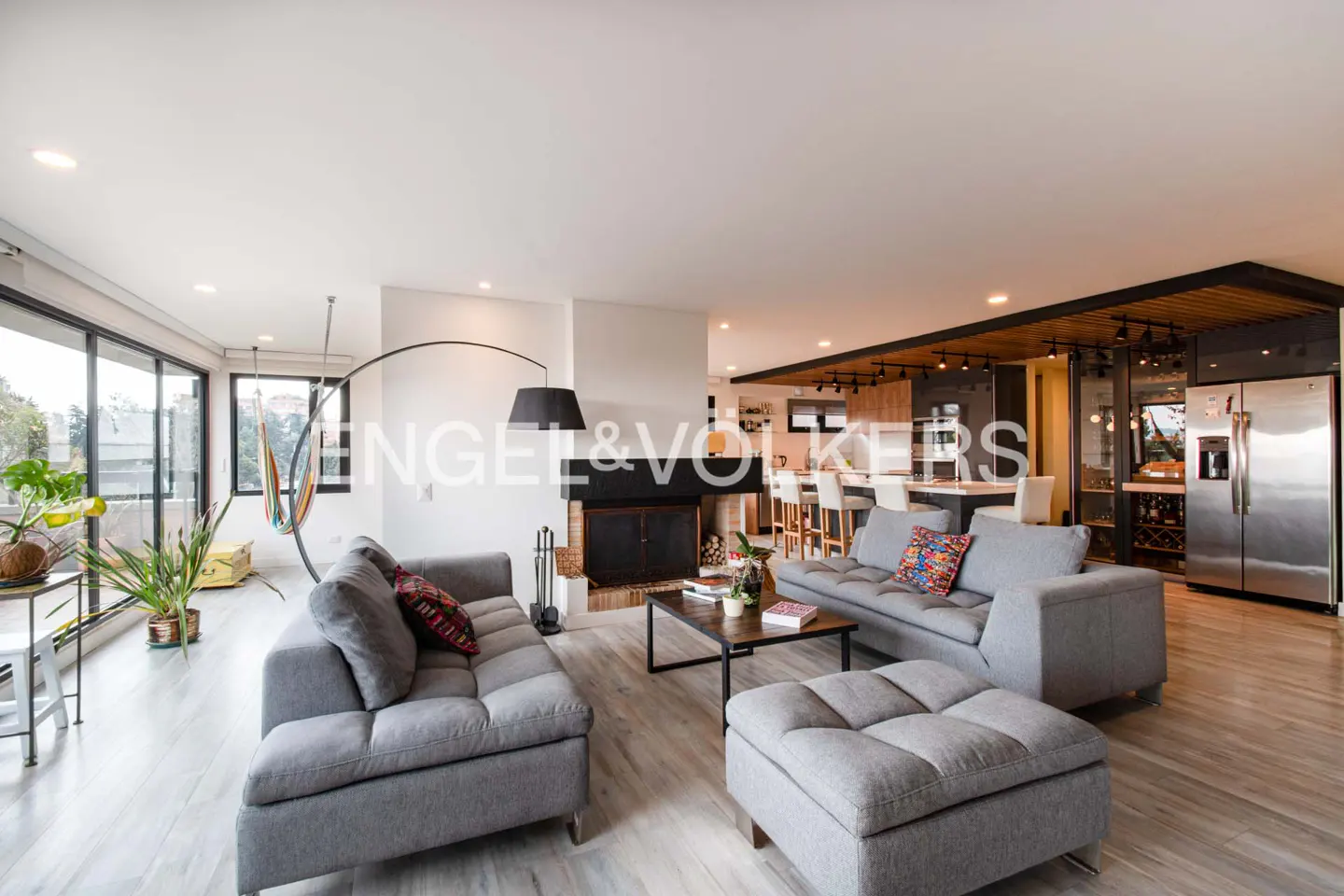 Open-concept living space with gray sofas, a fireplace, and a kitchen with a breakfast bar. Large windows provide natural light.