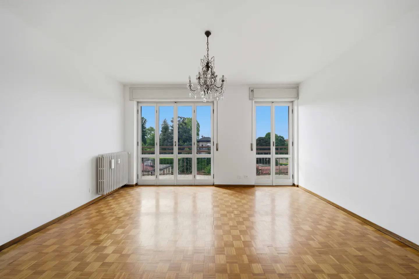 Empty white room with parquet floor, chandelier, radiator, and two sets of white French doors leading to balconies with green trees.