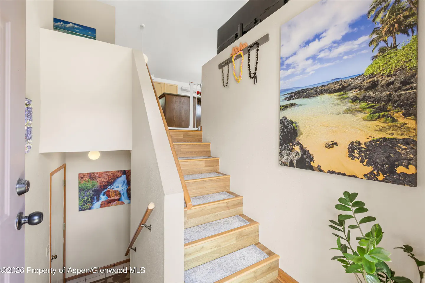 Entryway with wooden stairs and beach-themed decor. A tropical beach painting hangs on the wall. Leis are displayed above the stairs.