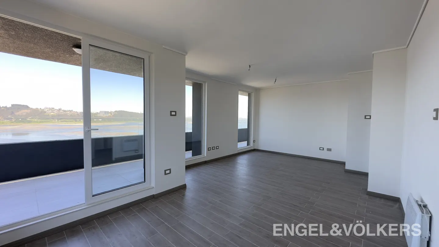 Bright, empty room with gray wood floors, white walls, and large windows overlooking a bay. Engel & Völkers logo in the corner.