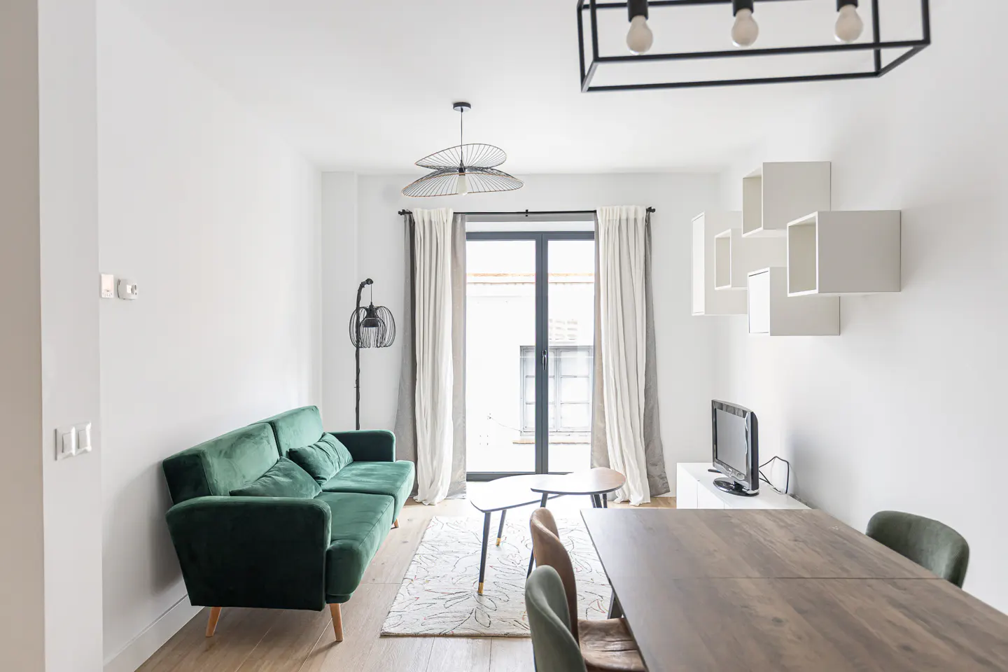 Bright living room with a green velvet sofa, wooden dining table, and white walls. A black framed window lets in natural light.