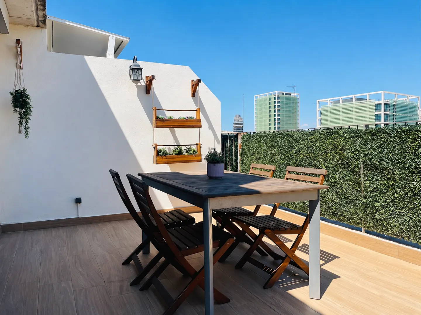 Outdoor patio with a dark wood table and chairs, a white wall with hanging plants, and a green hedge with buildings in the background.