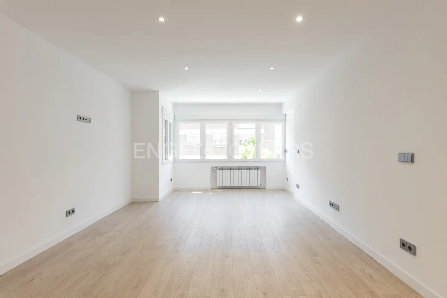 Bright, empty room with light wood floors, white walls, and a large window with a radiator.