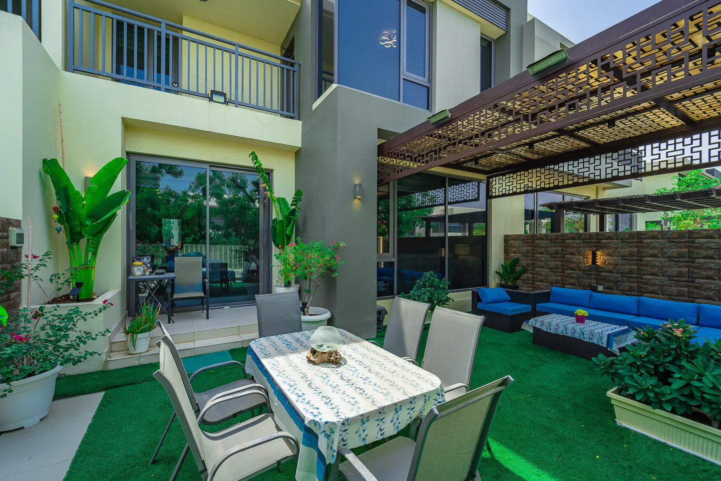Outdoor patio with a table and chairs on green turf, a blue sofa under a decorative pergola, and a modern building in the background.