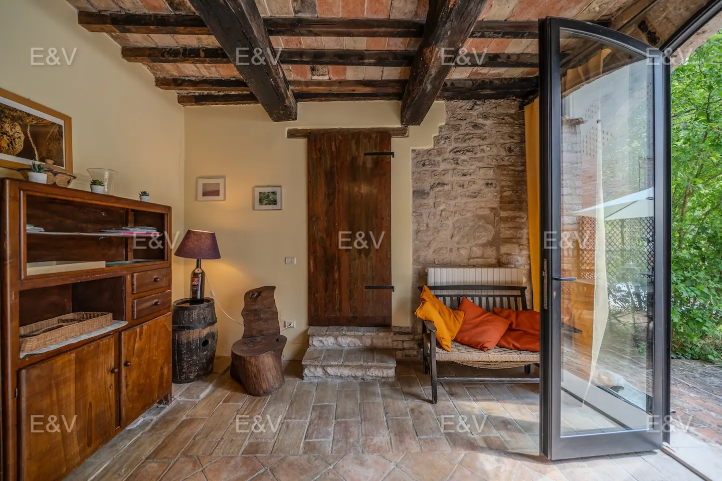 Rustic room with brick floors, wooden beams, and stone walls. A bench with orange pillows sits near an open glass door to a green outdoor space.