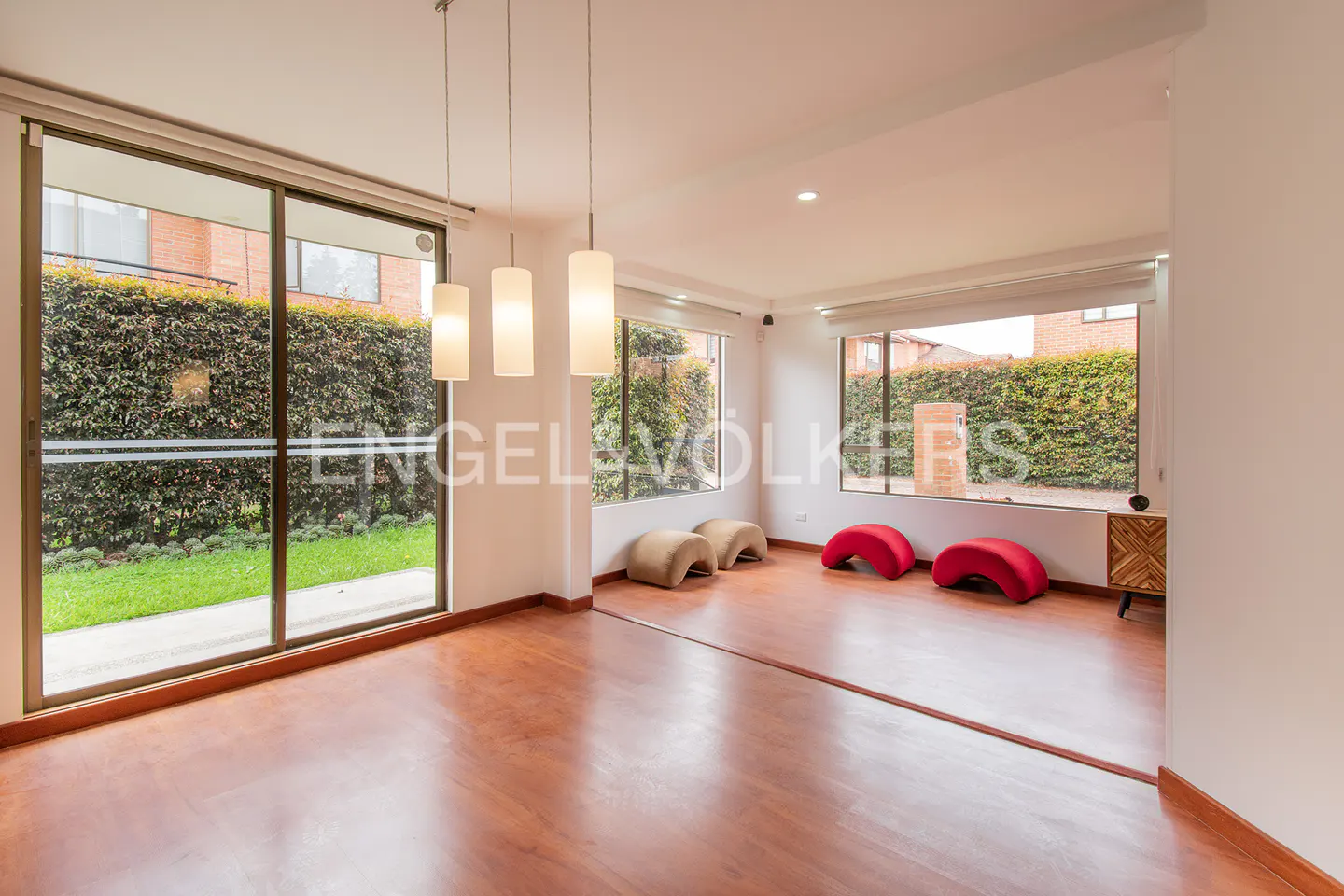 Bright living room with wood floors, large windows overlooking green hedges, and modern pendant lights. Red and beige seating.