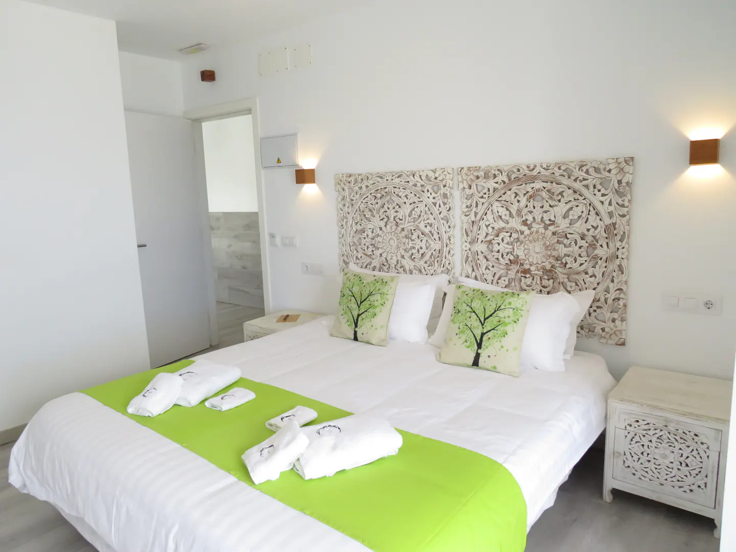 Bright bedroom with white walls, carved headboard, and green accents. Bed has a green runner and tree-patterned pillows. Nightstands with carved details.