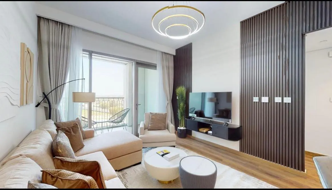 Living room with beige sectional sofa, round coffee tables, TV on a wood-paneled wall, and balcony with city view.