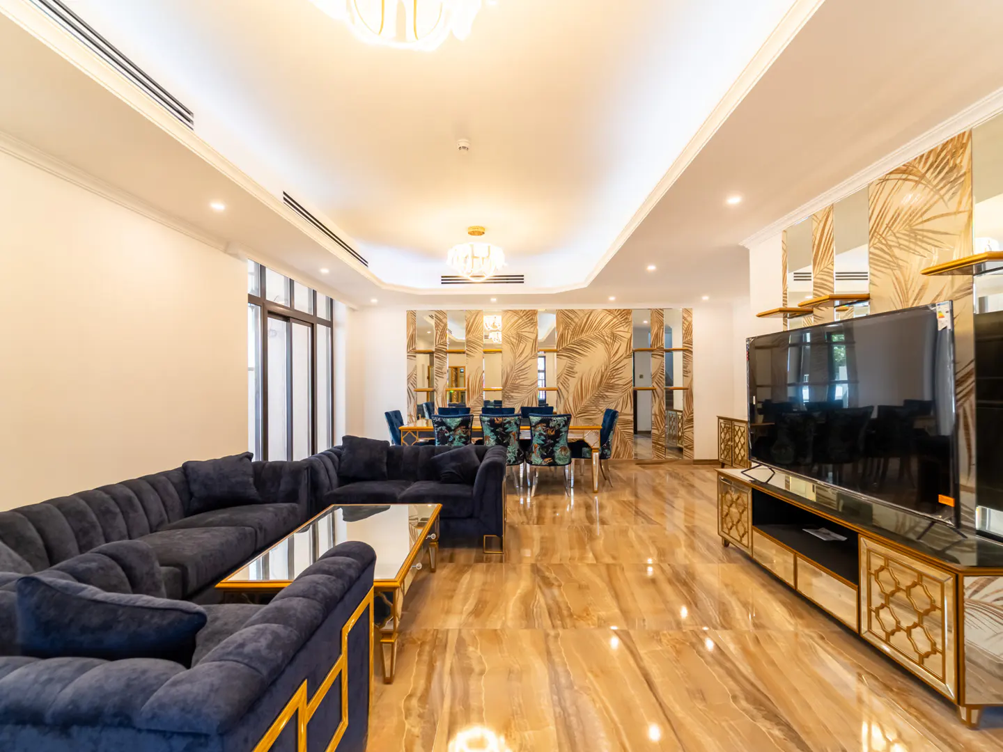 Modern living room with a navy blue sectional sofa, glass coffee table, large TV, and dining area with palm-patterned walls.