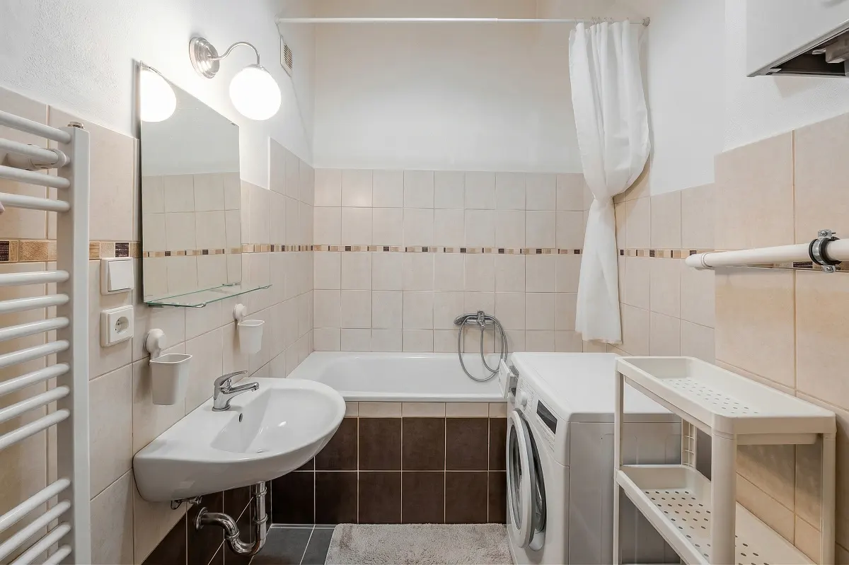Small bathroom with beige and brown tiles, featuring a white sink, bathtub, washing machine, and a white storage shelf.