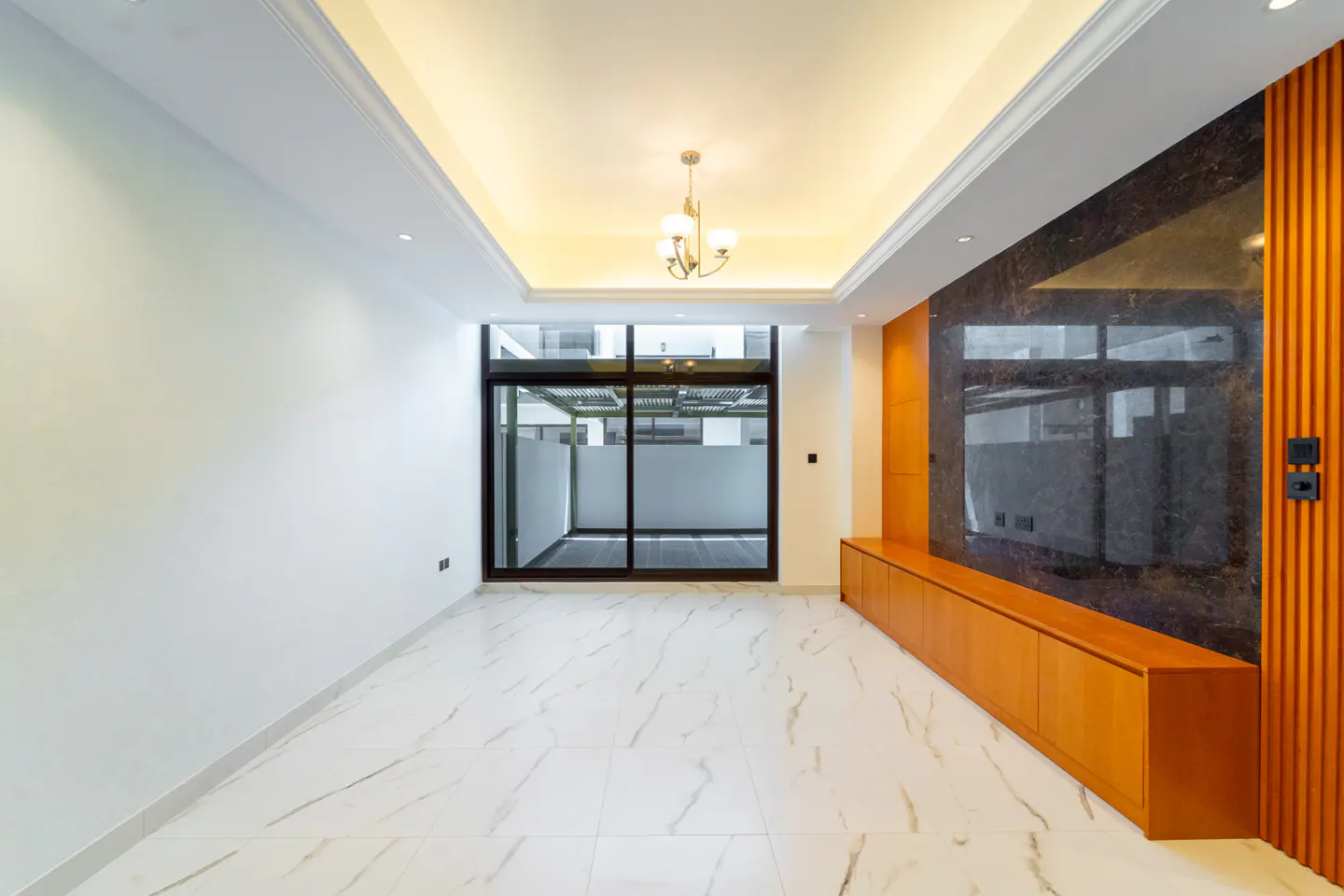 Bright, empty room with white walls, marble floors, and a brown cabinet. A sliding glass door leads to a patio. A chandelier hangs from the ceiling.