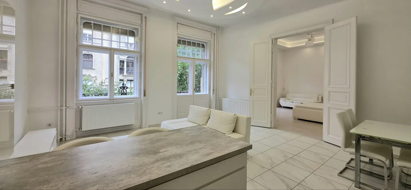 Bright, modern living room with white walls, tile floors, large windows, and a view into an adjacent room with a sofa.