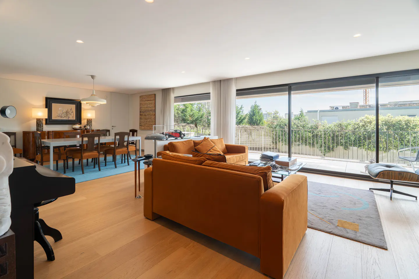 Bright living room with wood floors, orange sofas, dining table, and large windows overlooking greenery.