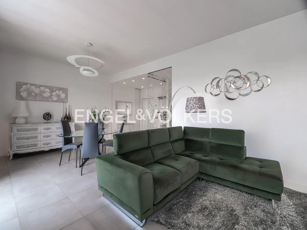 A modern living room with a green sectional sofa, gray rug, and dining area with a glass table. White walls and a decorative metal wall sculpture.