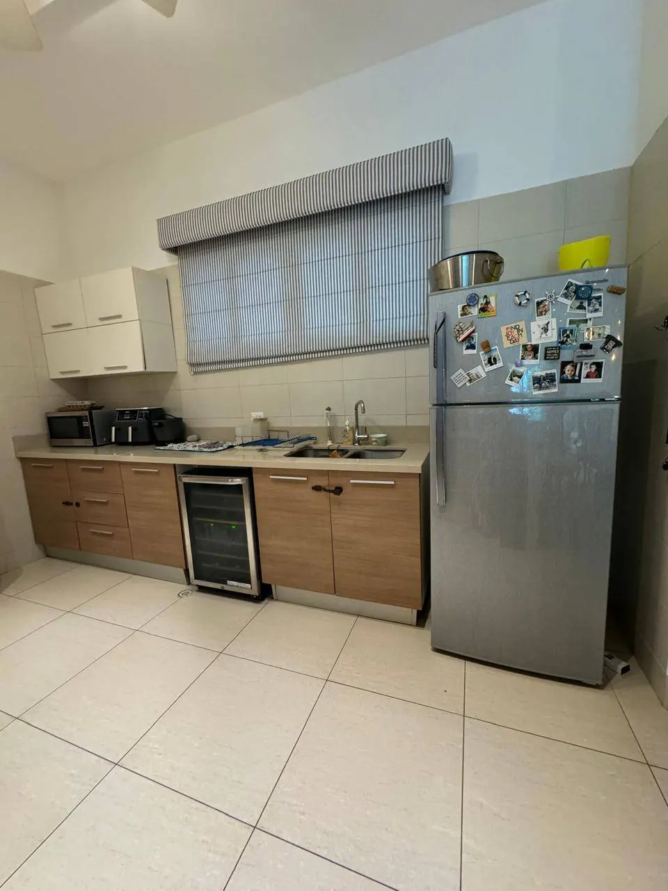 A kitchen with light wood cabinets, white countertops, and a stainless steel refrigerator covered in magnets. A striped blind covers the window.