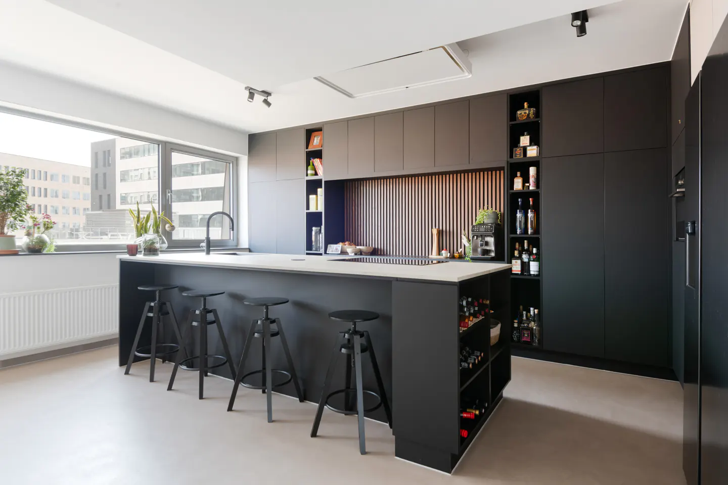 Modern kitchen with a black island, white countertop, and four black stools. Black cabinets line the wall with open shelving and a wine rack.