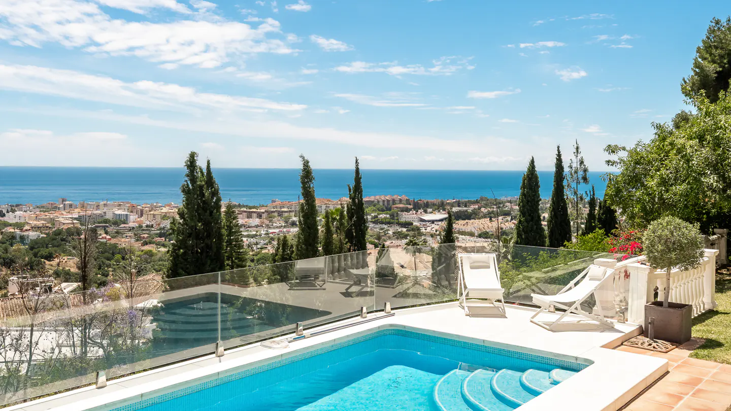 A blue tiled pool with steps, two white lounge chairs, and a glass fence overlook a city and the ocean under a blue sky.