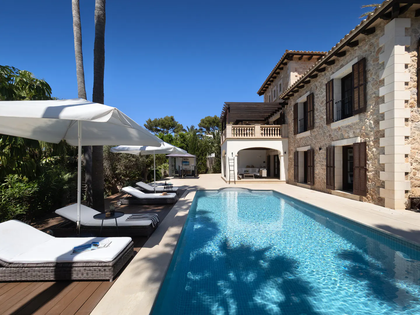 Exterior view of a stone house with a pool, lounge chairs, and white umbrellas on a sunny day.