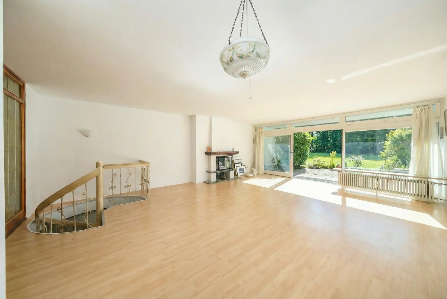Bright, empty living room with wood floors, white walls, and a spiral staircase. A fireplace and large windows overlook a green yard.
