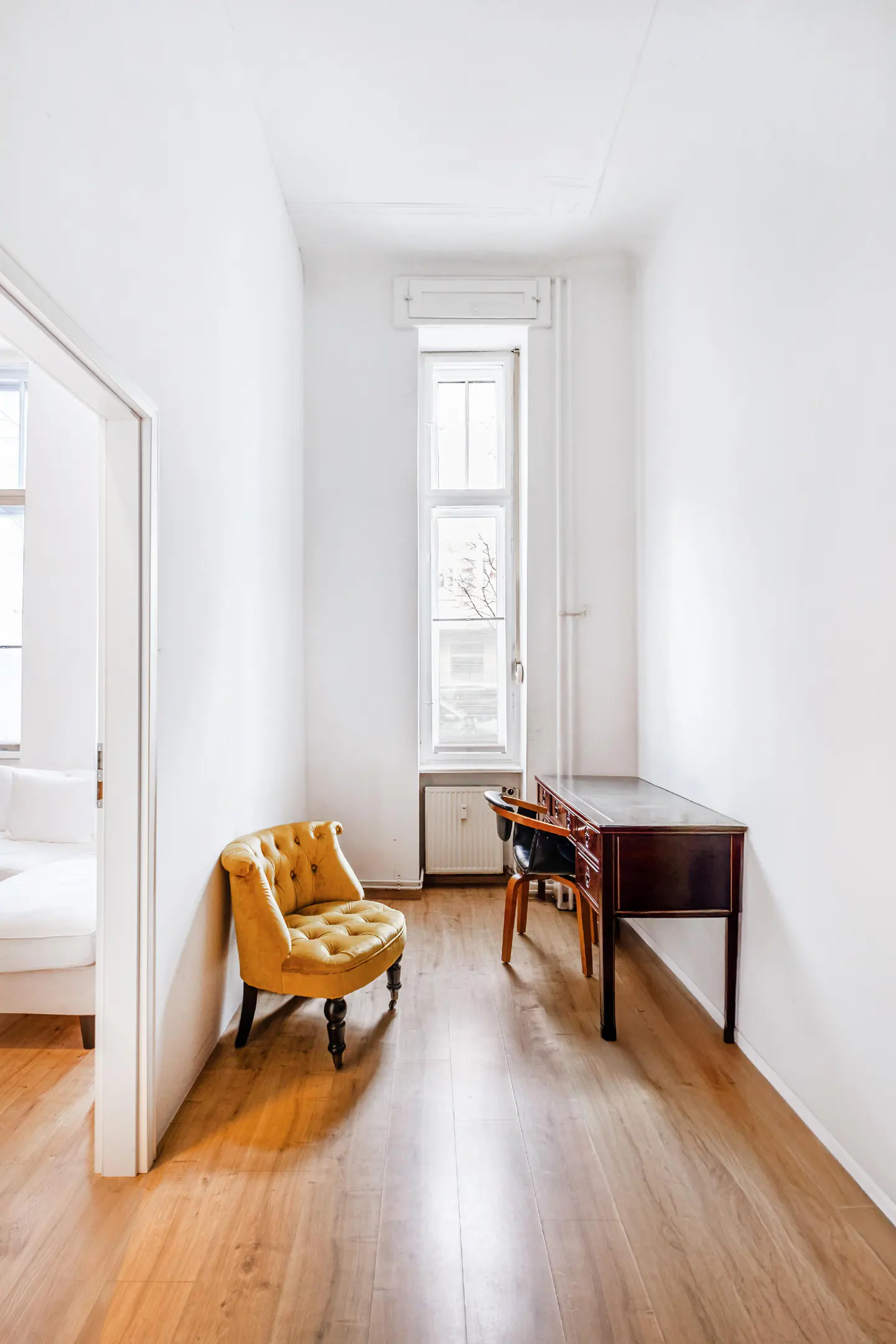 Bright room with wood floors, white walls, and a window. A yellow chaise lounge sits near an open doorway, and a dark wood desk with a chair is by the window.