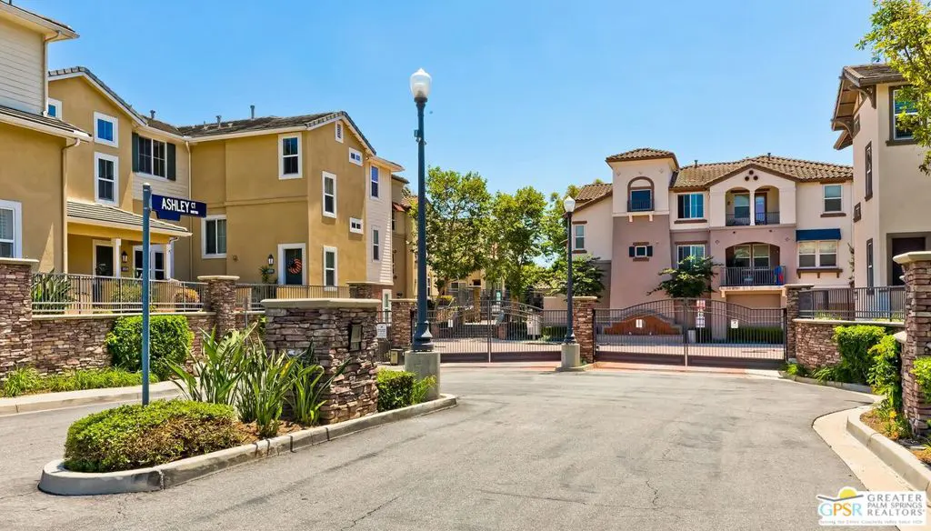 Exterior view of a gated community with tan and beige townhouses under a clear blue sky. "Ashley St" sign visible.
