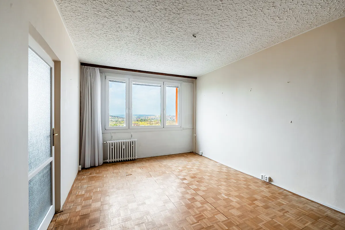 Empty room with parquet floor, white walls, and textured ceiling. A window with white curtains overlooks a cityscape. A white door is on the left.