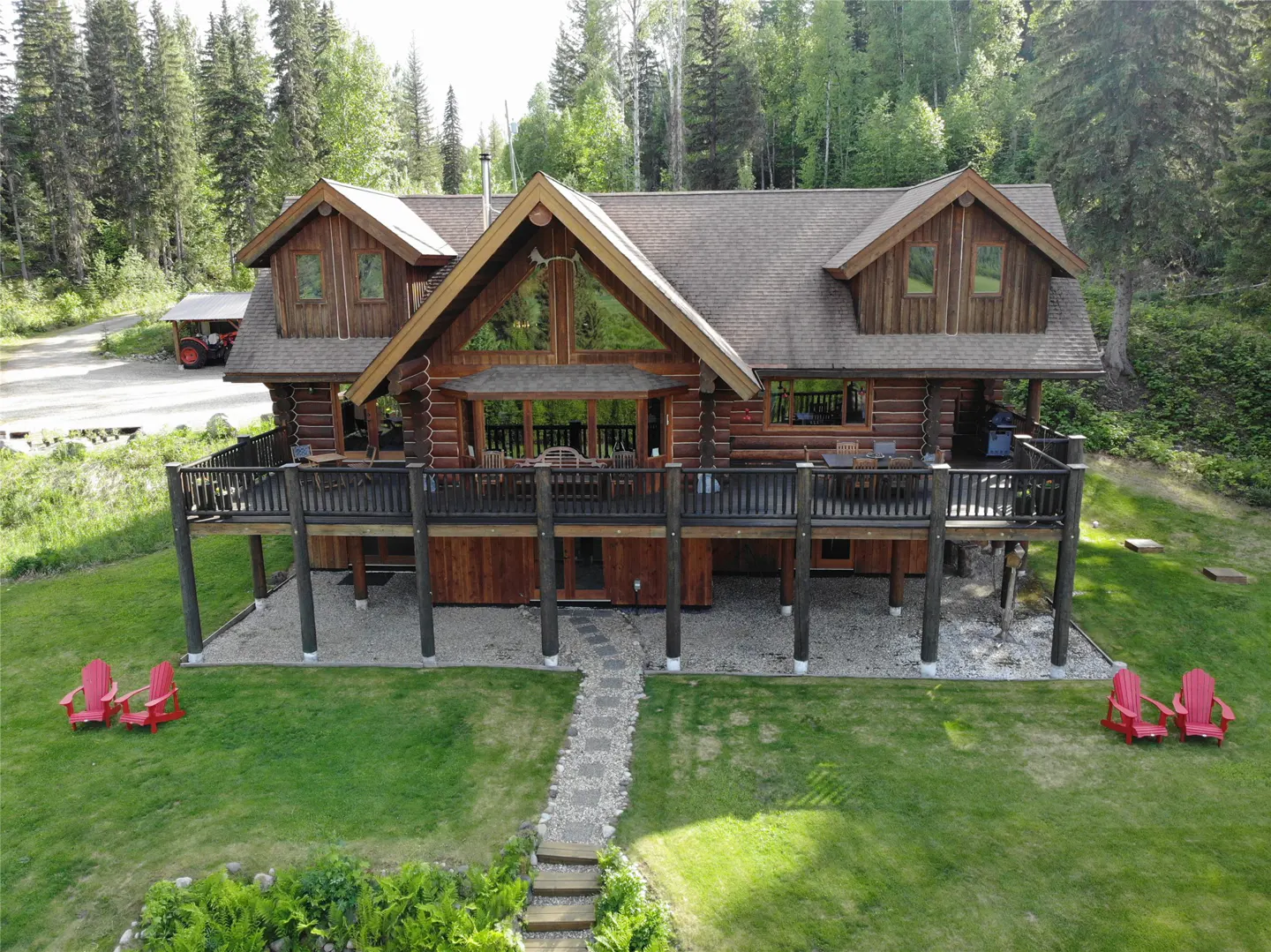 Aerial view of a log cabin home with a large deck, green lawn, stone path, and red Adirondack chairs.
