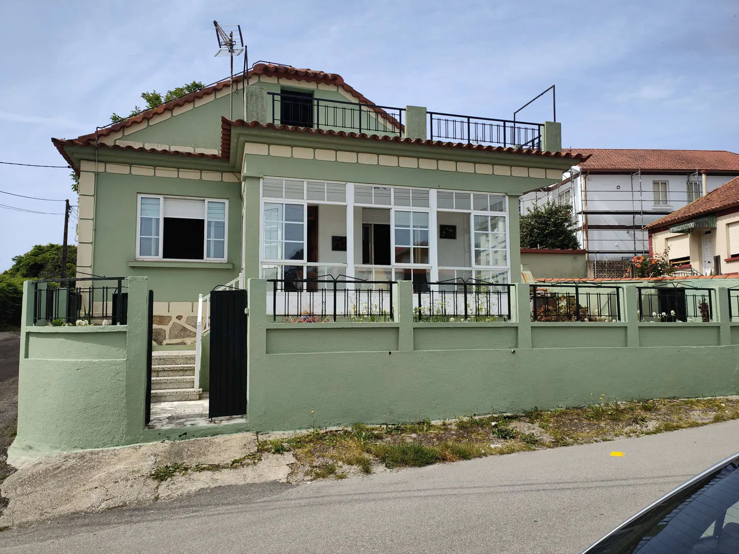 A light green house with a red tile roof and white-framed windows, surrounded by a low green wall with a black metal fence.