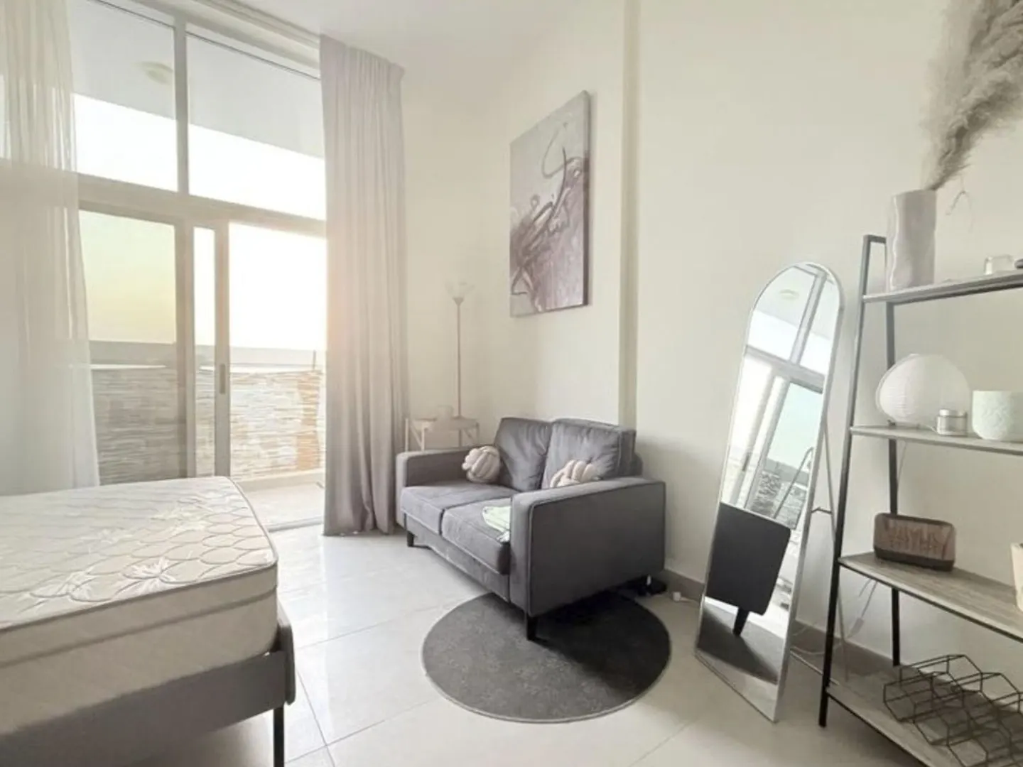 Bright, modern apartment interior with a gray sofa, bed, and shelving unit. A large window with sheer curtains lets in natural light.