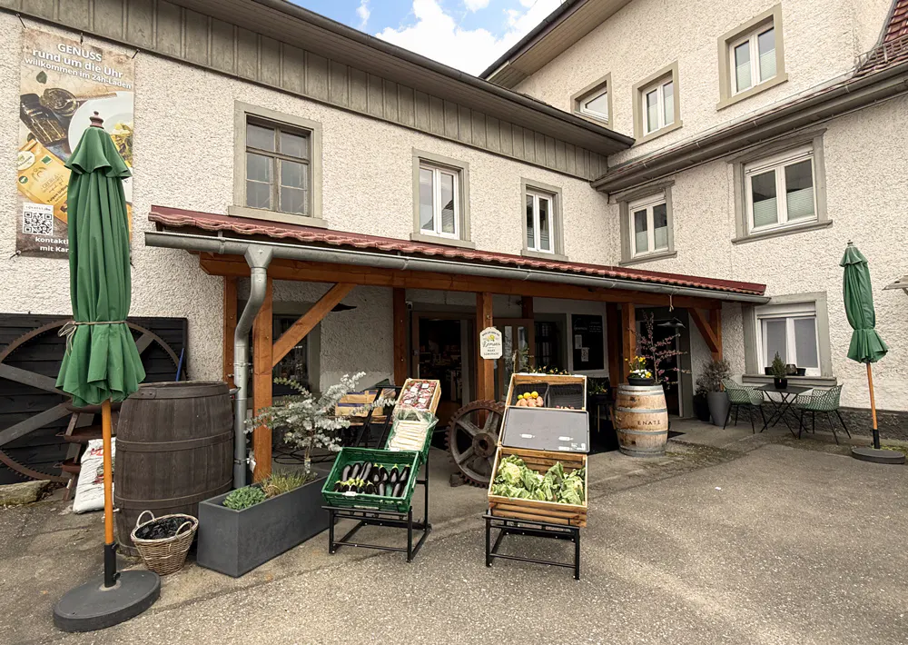 Exterior view of a two-story building with produce stands in front. Green umbrellas, barrels, and plants decorate the entrance.
