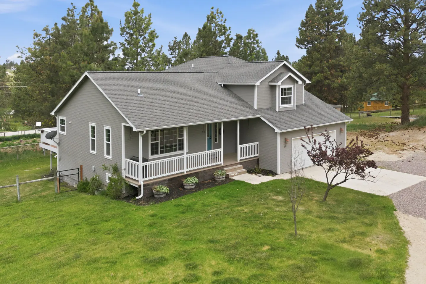 Two-story gray house with a gray roof, white porch railing, and a turquoise front door. Green lawn and trees surround the house.