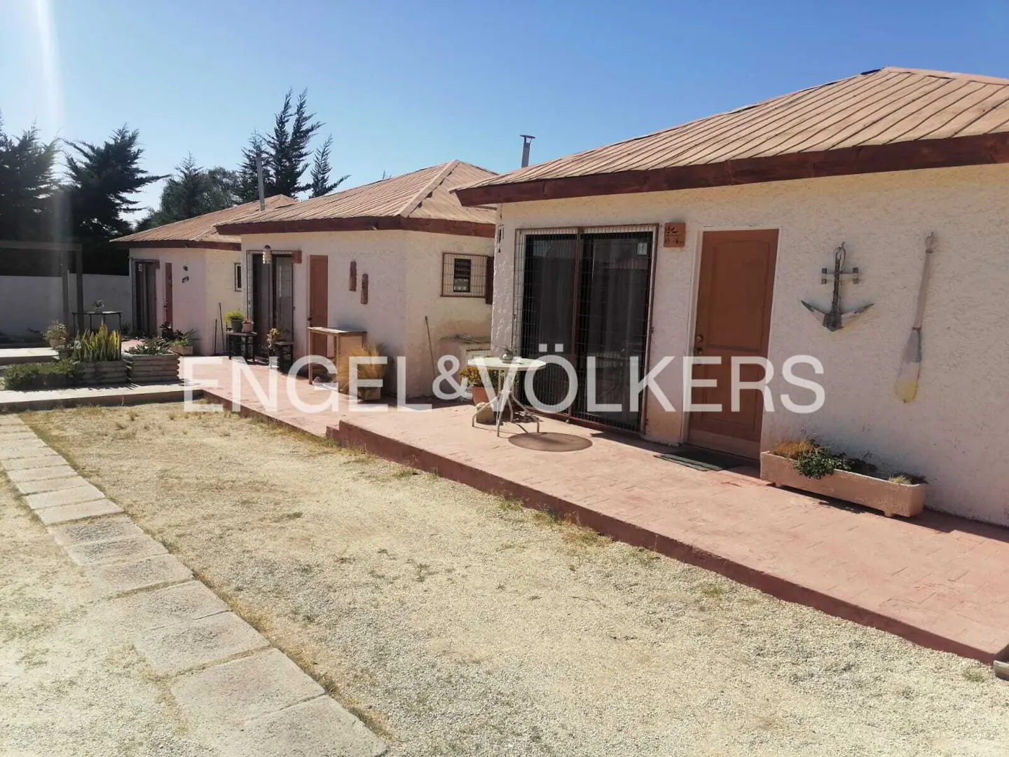 Row of small, beige stucco vacation cottages with brown tile roofs and a shared red-tiled patio under a clear blue sky.