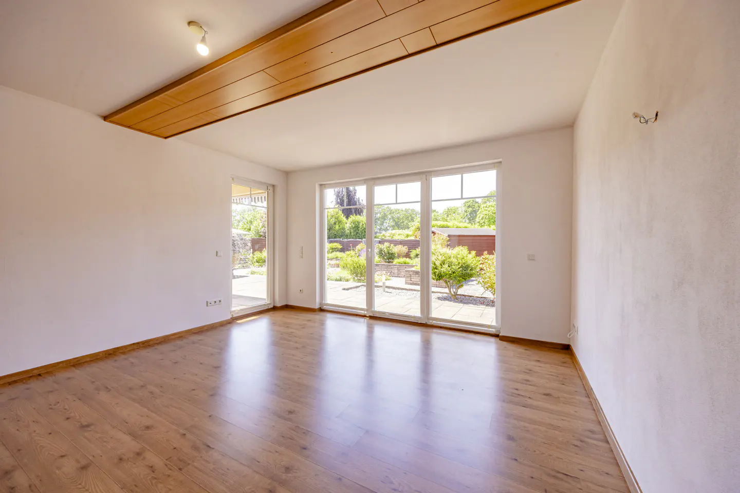 Empty room with light wood floors, white walls, a wooden ceiling beam, and large glass doors leading to a garden patio.