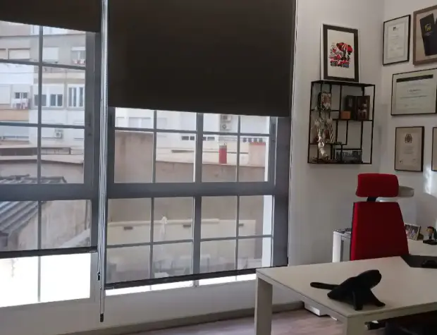 Office interior with large windows covered by dark roller shades. A white desk with a black cat figurine sits in front of a red office chair. Wall art and a shelf decorate the white wall.