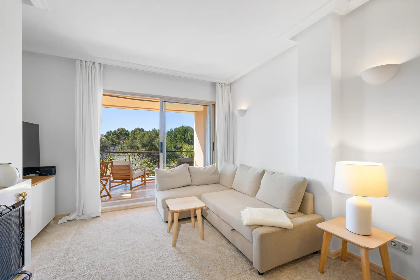 Bright living room with beige sectional sofa, wood side tables, and balcony with chairs overlooking green trees.