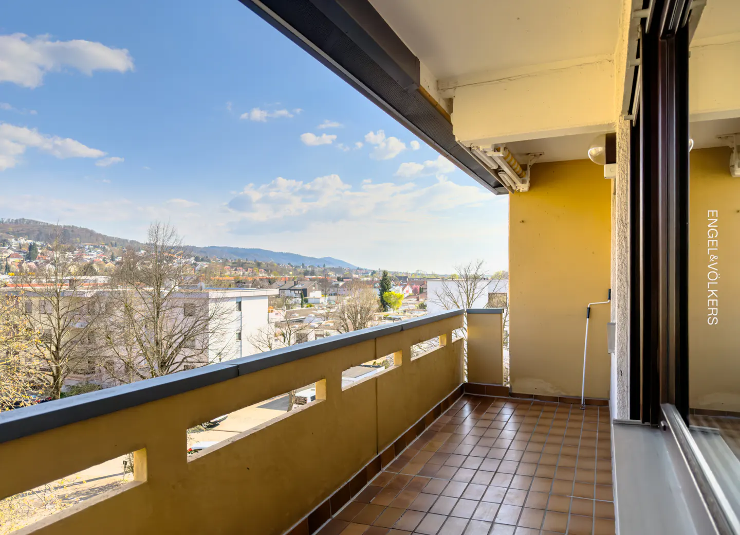 Balcony view of a city with a blue sky. The balcony has brown tile and a yellow railing with rectangular cutouts.
