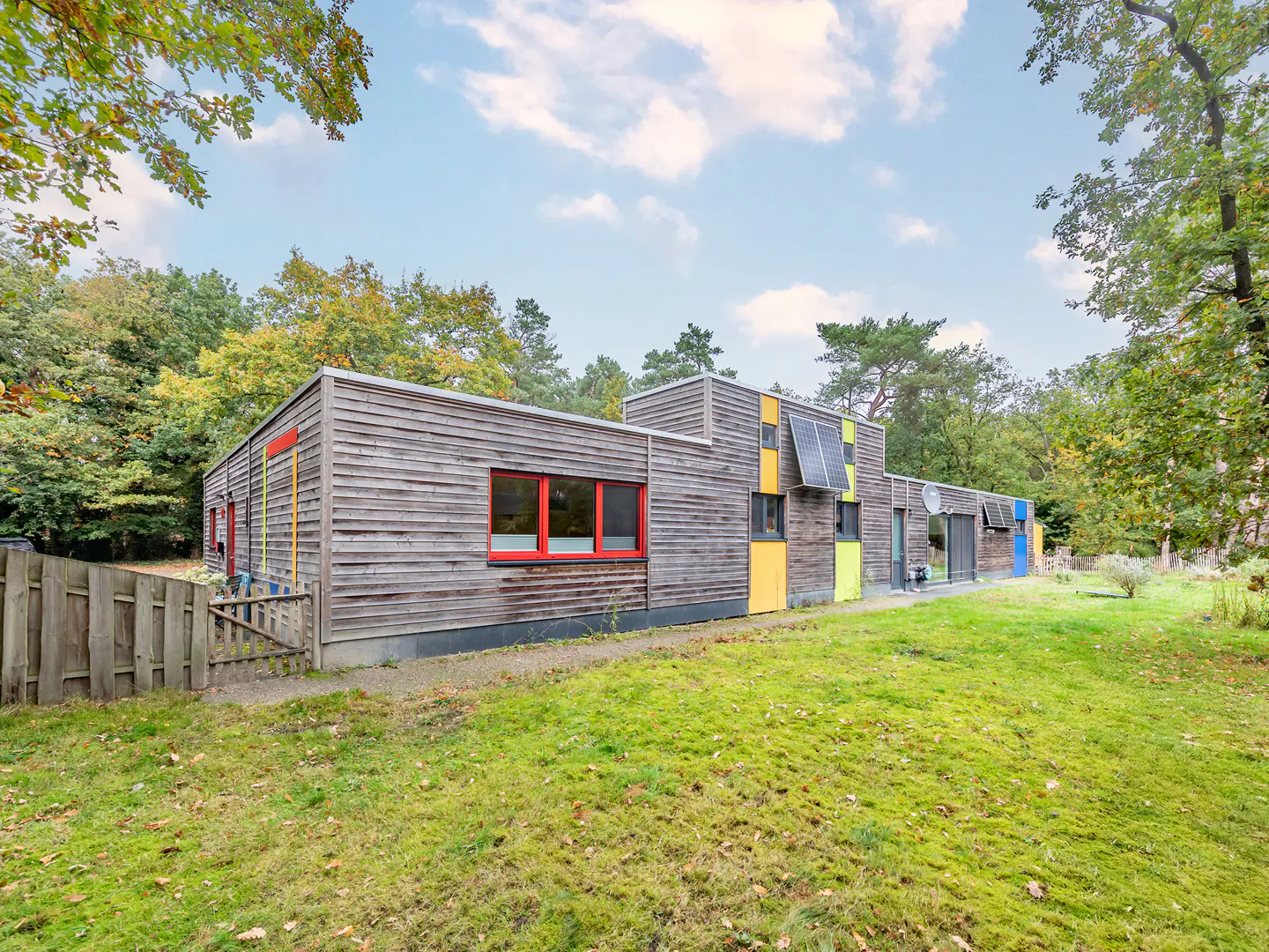 Exterior view of a modern, wood-sided house with colorful window accents and solar panels, surrounded by green lawn and trees.