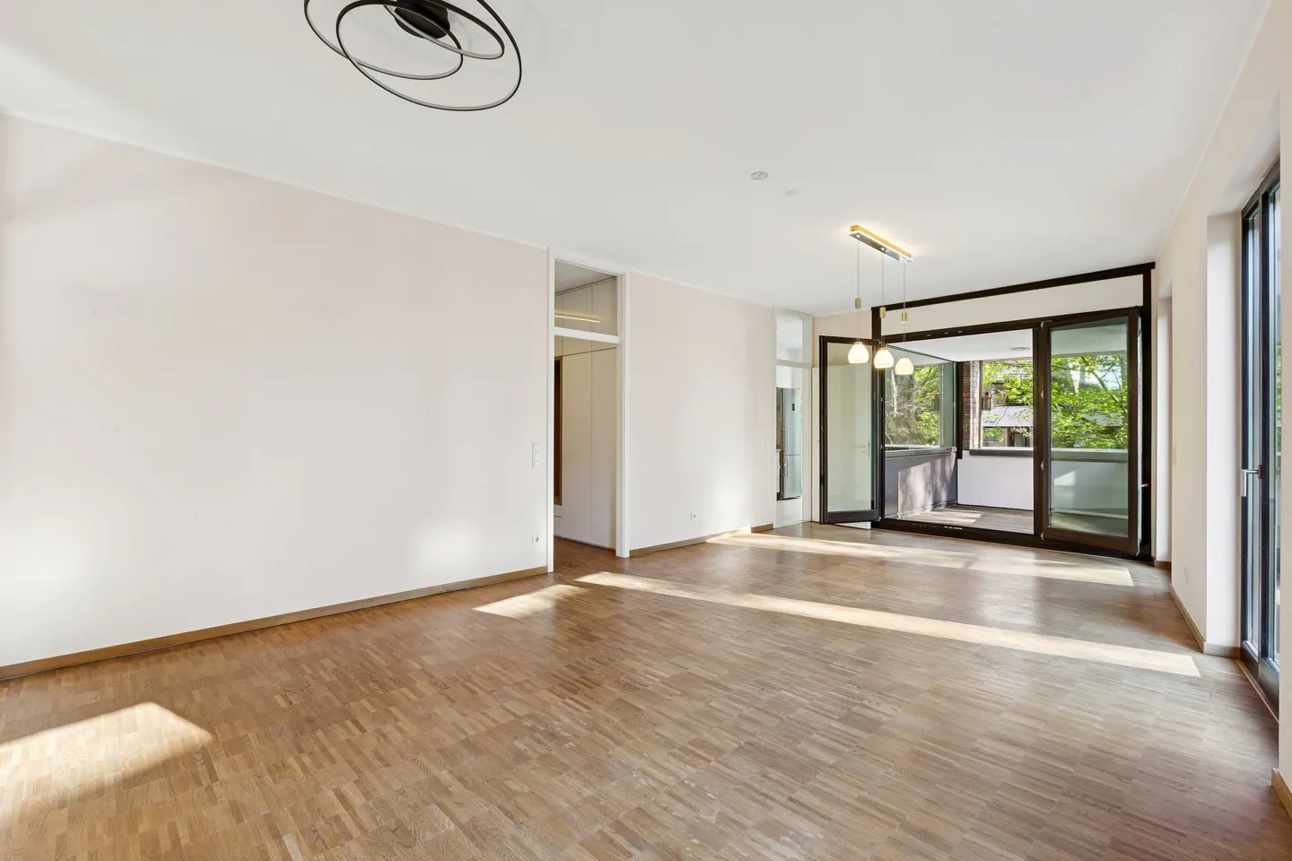 Bright, empty room with wood floors, white walls, and black framed glass doors to a balcony. Sunlight streams in.
