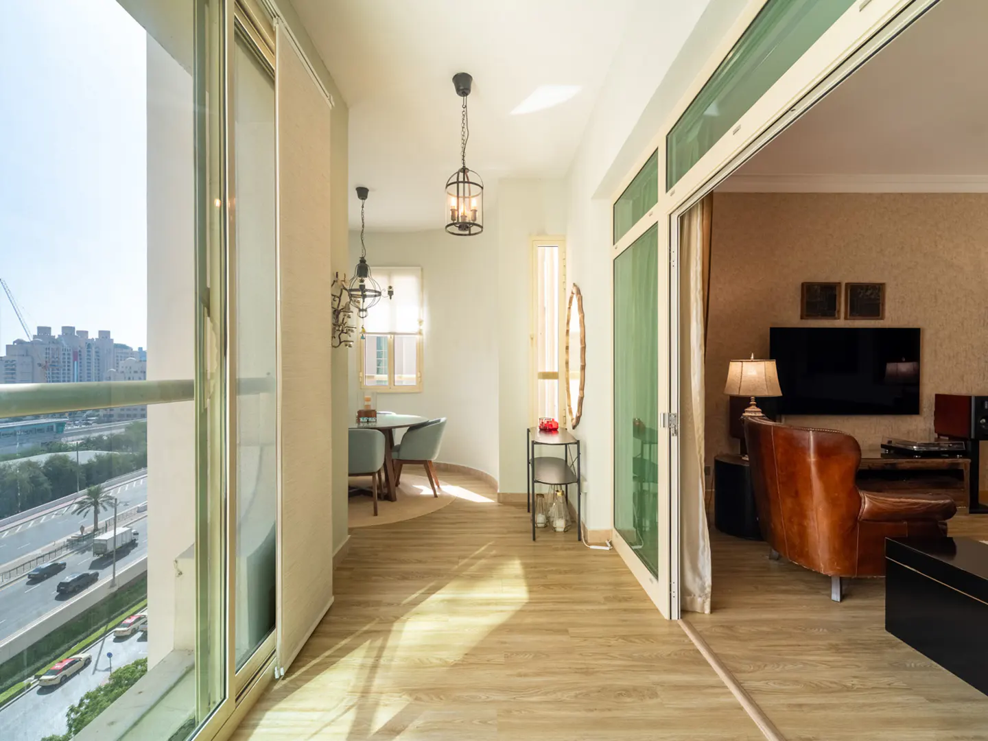 Bright balcony with wood floors, hanging lanterns, and a view of a city road, leading into a cozy living room.
