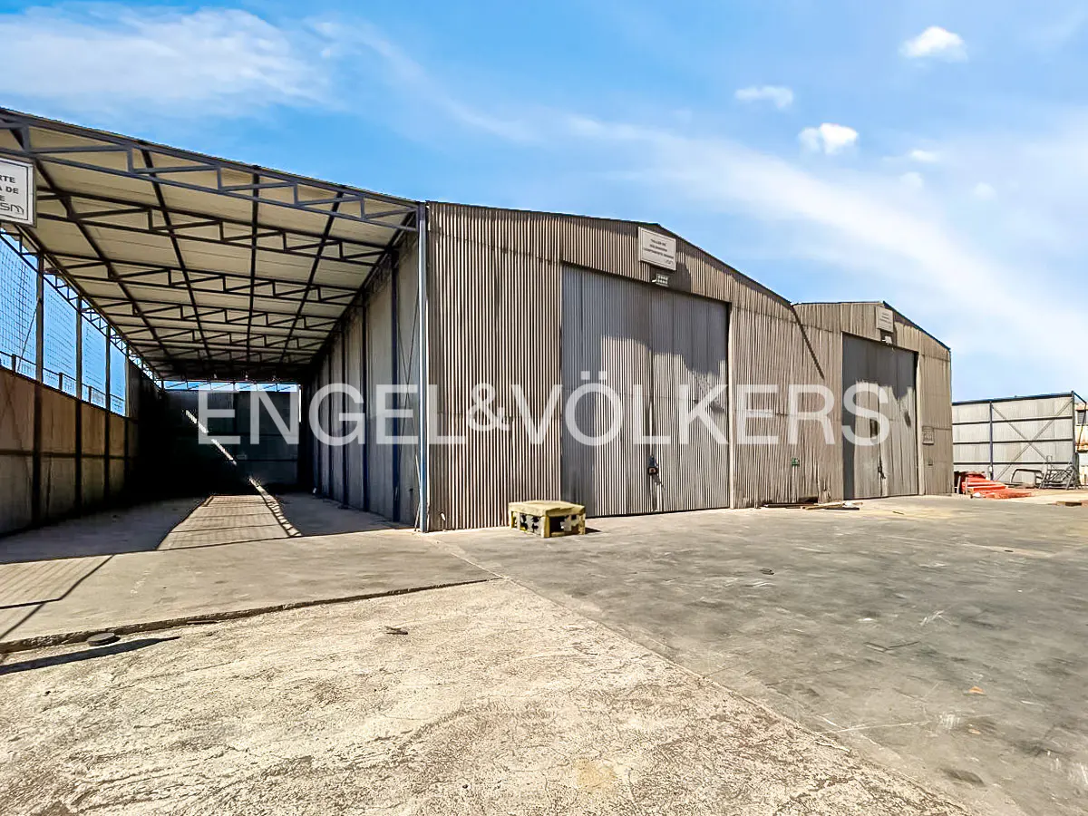 Exterior view of a large, gray metal warehouse with loading docks and the Engel & Völkers logo. Blue sky background.