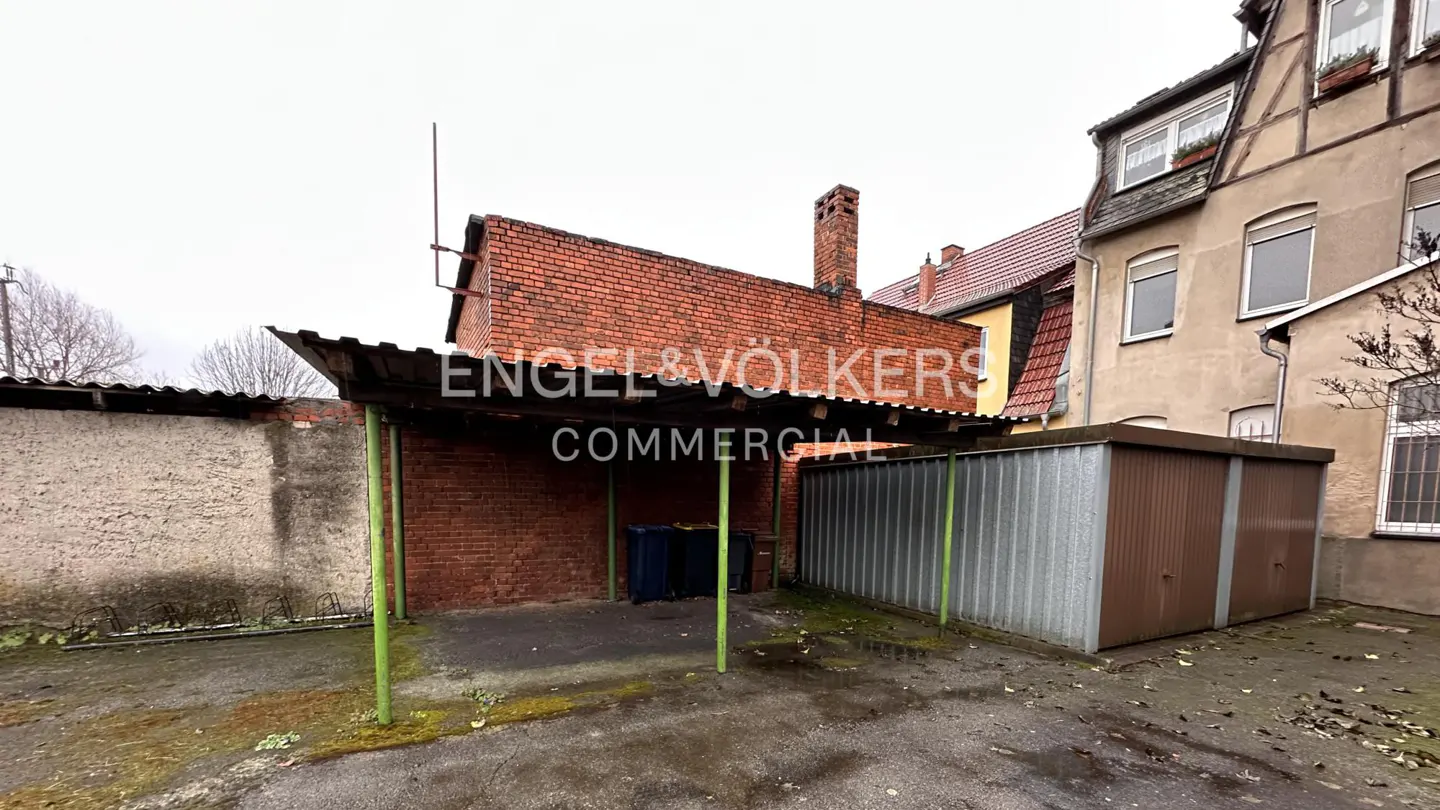 Outdoor shot of a carport with green supports, a brick wall, and a metal garage, with the Engel & Völkers logo.