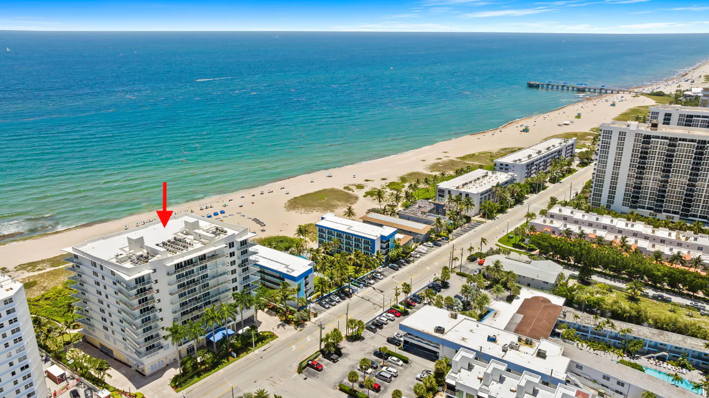 Aerial view of a white condo building on a beach, with an arrow pointing to the top. Blue ocean and sky in the background.