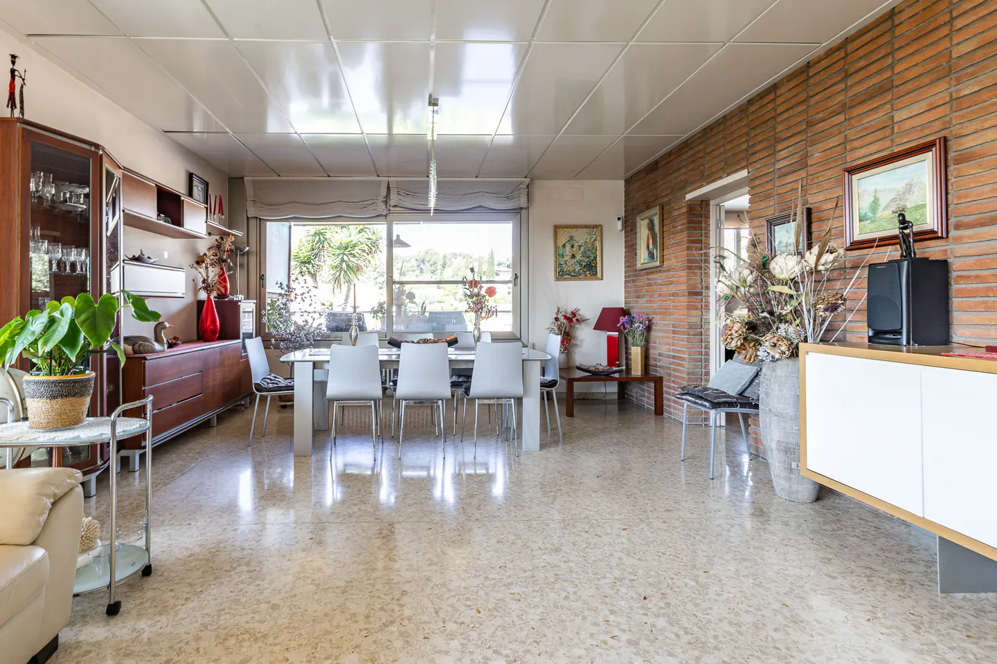 A bright dining room with a white table, six chairs, and a brick accent wall. Large windows offer a view of greenery.