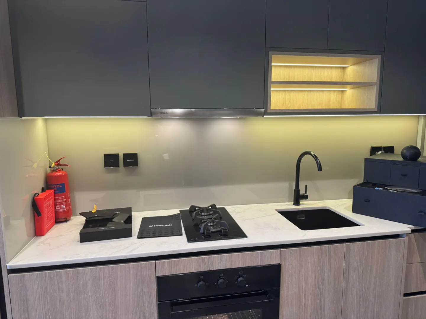 Modern kitchen with gray cabinets, black sink and faucet, white countertop, and wood-look lower cabinets. A red fire extinguisher is visible.