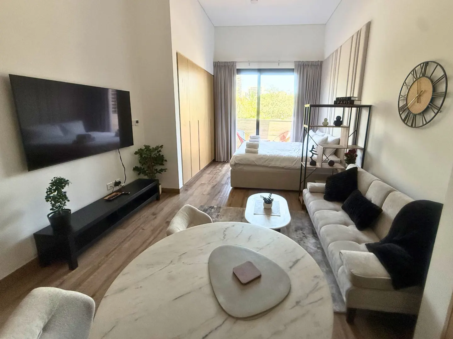A modern studio apartment with a bed, sofa, TV, and round marble table. The room has light wood floors and neutral-colored walls.
