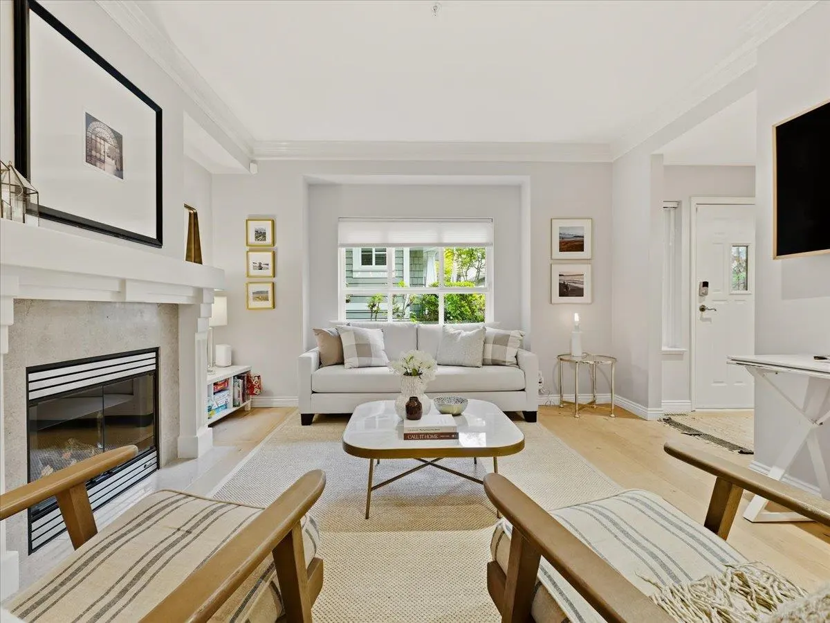 Bright living room with a white sofa, marble coffee table, fireplace, and two striped armchairs. A large window overlooks a green yard.