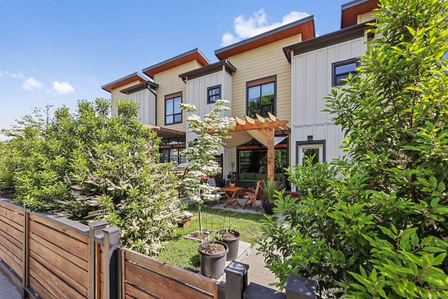 Modern townhouse exterior with a private patio, wooden pergola, lush landscaping, and a wooden fence under a blue sky.