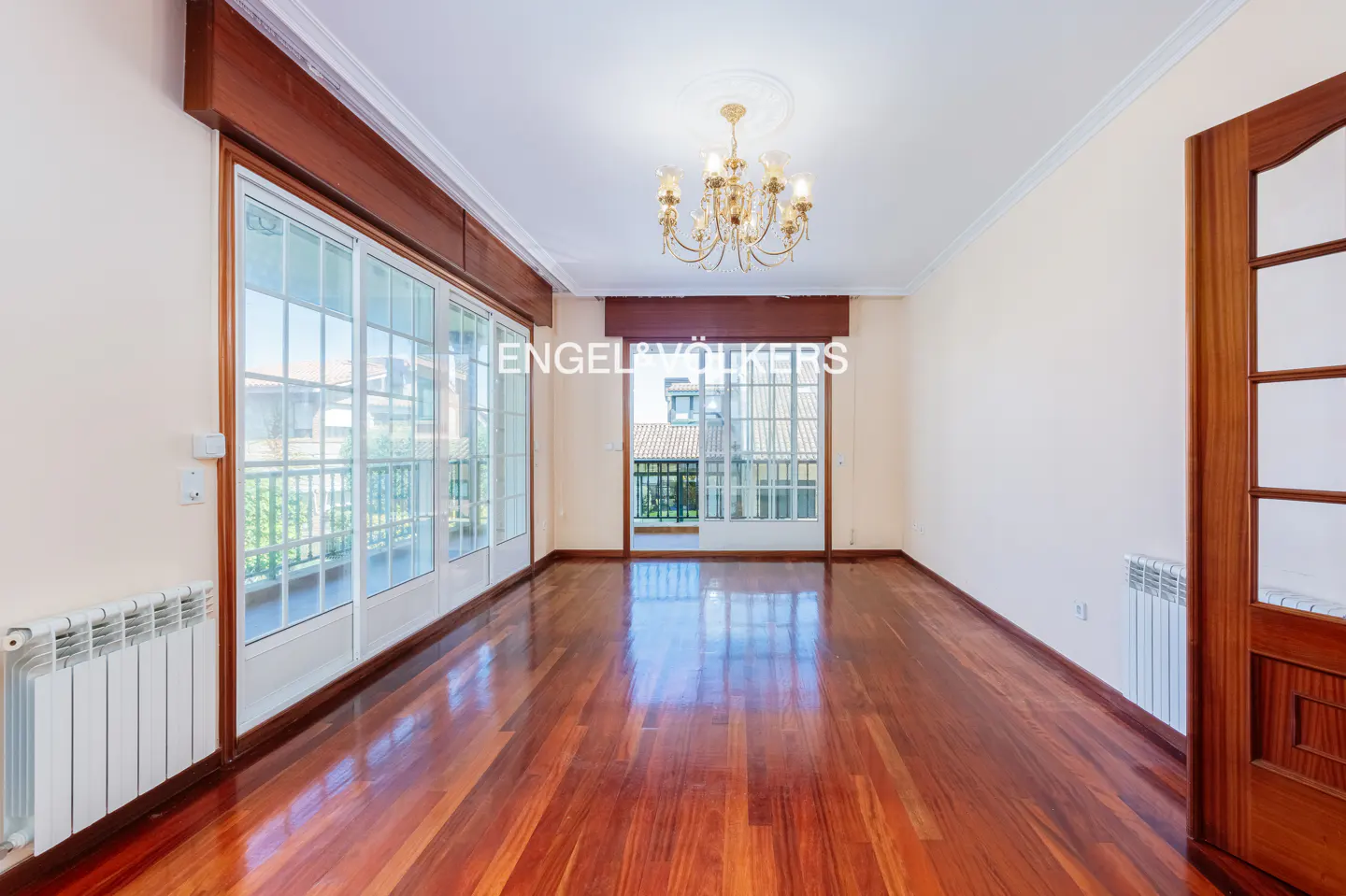 Empty room with polished hardwood floors, white walls, a crystal chandelier, and large glass doors leading to a balcony.