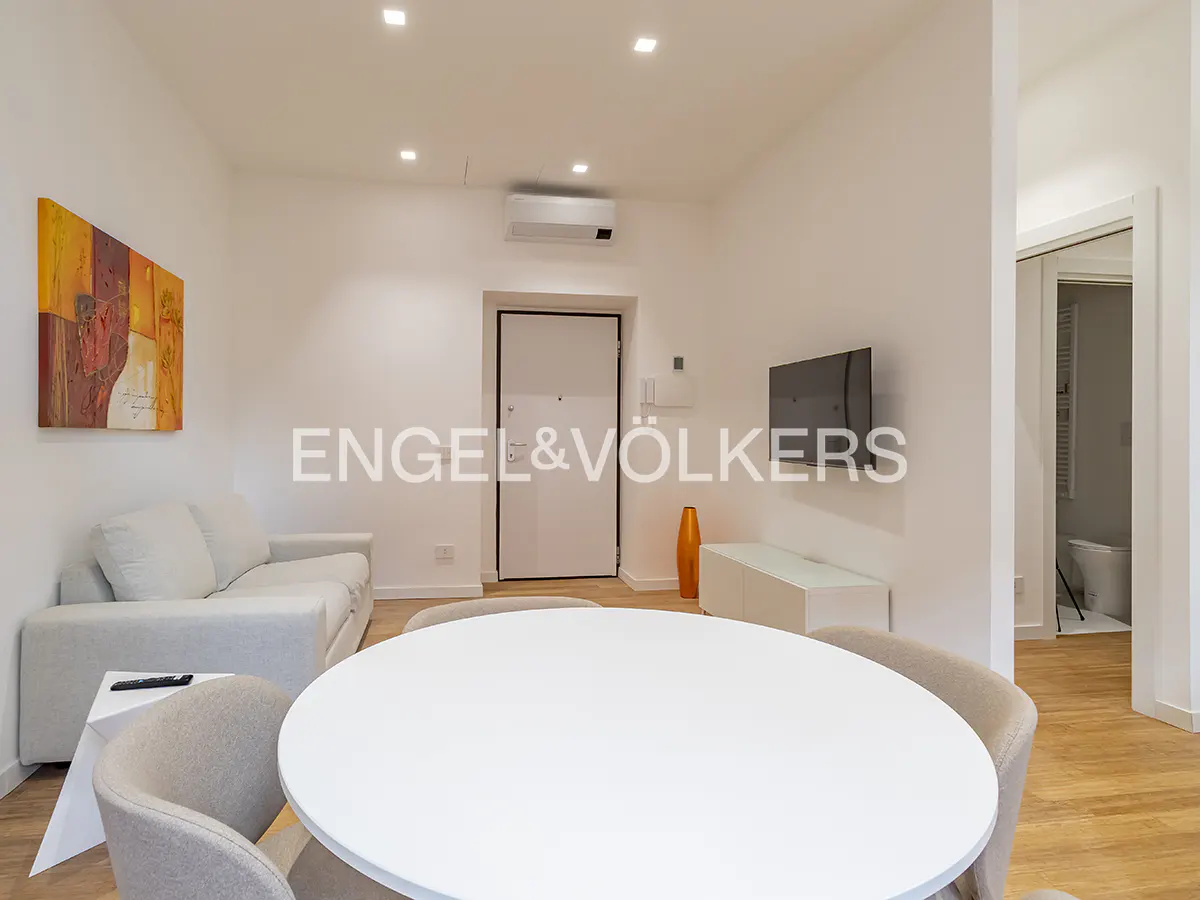 Bright living room with white walls, wood floors, and modern furniture. A round white table is in the foreground. A gray sofa and colorful art are visible.